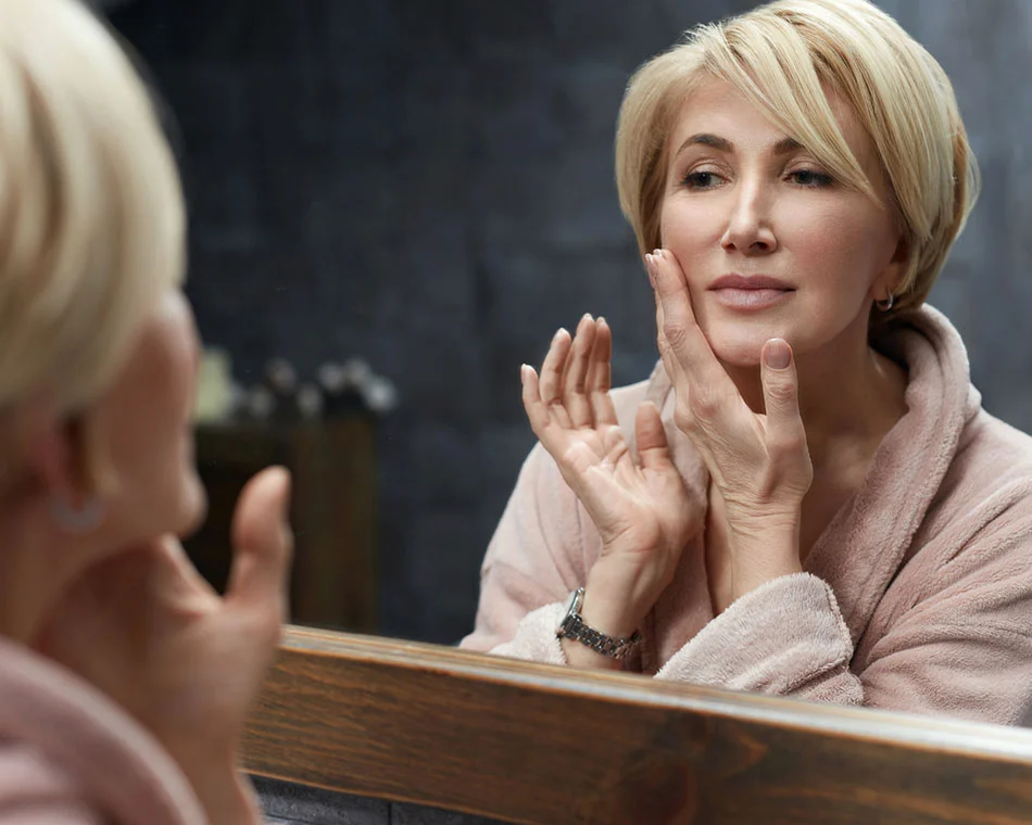 A woman in a bathrobe gently touches her face, admiring her reflection in a mirror, surrounded by a softly lit background. - Buccal Fat Pad Removal in Scottsdale, AZ