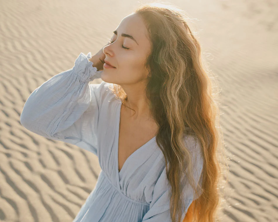 A woman with long, wavy hair stands in a sunlit desert, eyes closed, enjoying the warm breeze, wearing a light blue dress. - Upper Eyelid Surgery in Scottsdale, AZ